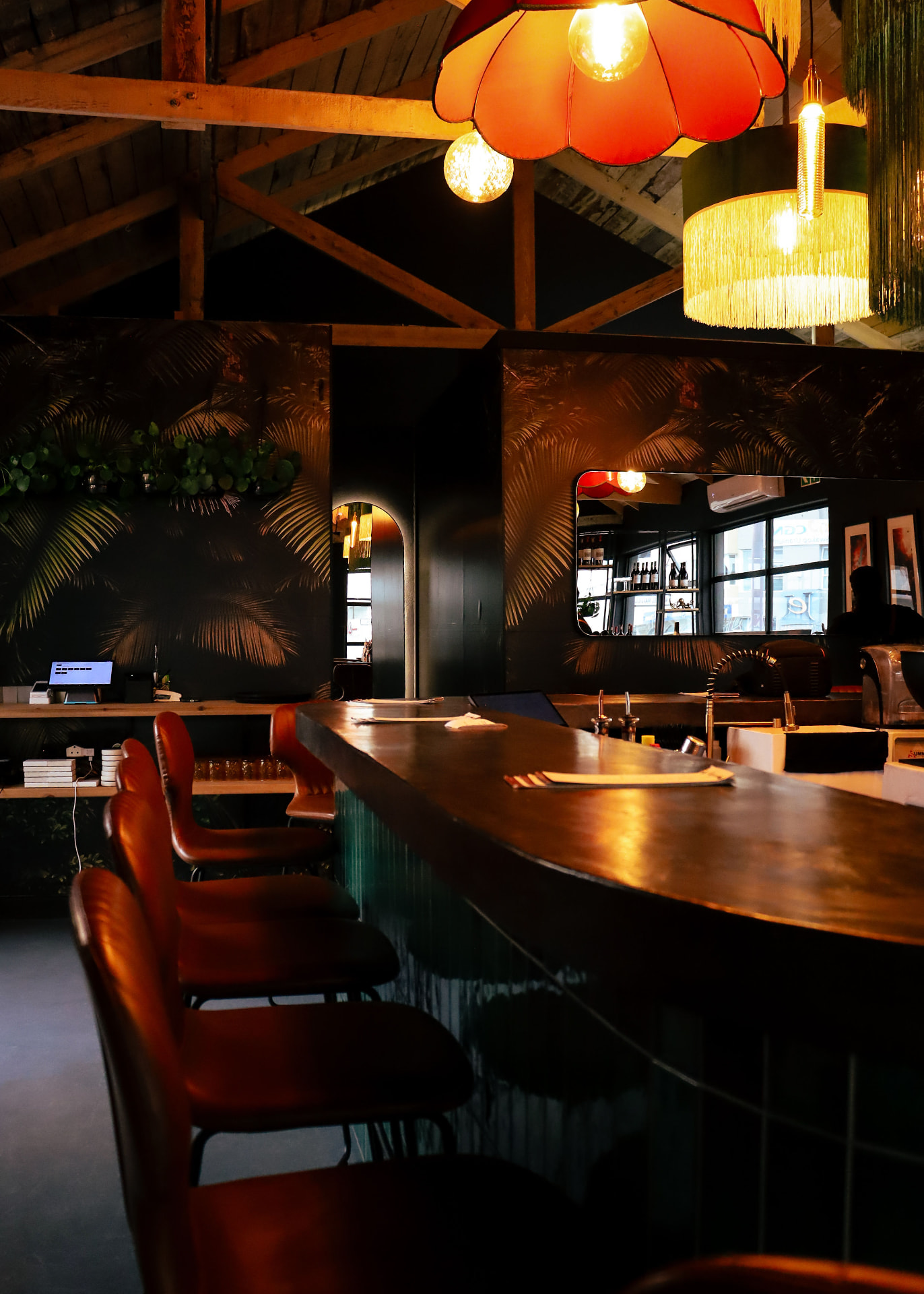 The curved bar counter under fringed pendant lamps and painted palm walls